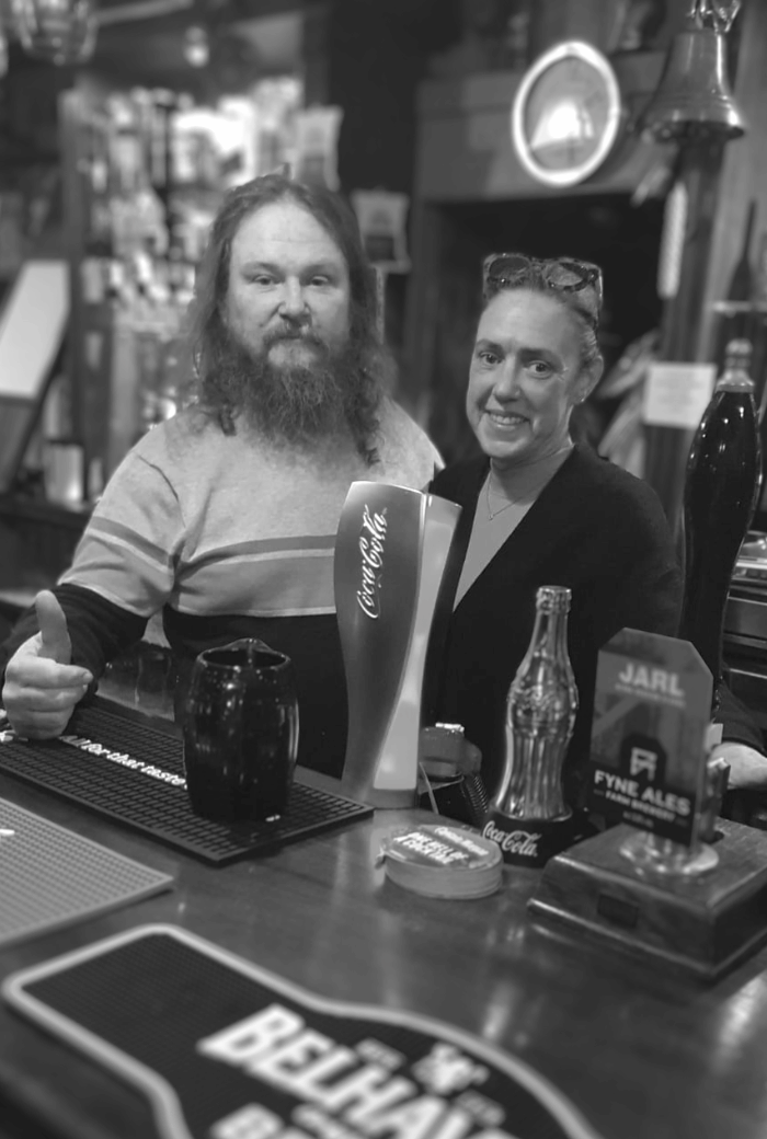 Inside The Clachnaharry Inn, a man and a woman stand behind the bar, smiling amidst an array of Scottish whiskies and real ales, including Belhaven Best and Fyne Ales, symbolizing the pub's rich tradition and family-run warmth